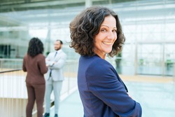 Happy successful business woman posing with arms folded, looking at camera, smiling. Her male and female colleagues standing and talking in background. Confident business leader concept