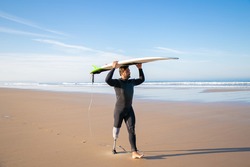 Male surfer wearing artificial limb and wetsuit, walking on beach, carrying surfboard overhead. Full length. Artificial limb and active lifestyle concept