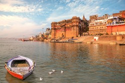 Ancient Varanasi India city architecture with view of Ganges river ghat at sunset. Wooden boat seen on river Ganga with migratory birds