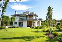 Modern country house with the large lawn and a wooden fence. In front of the house there is a covered terrace with a lounge zone. On the lawn there is a trees and flowerbeds. It is sunny.