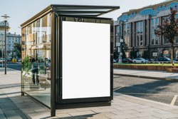 Vertical blank white billboard at bus stop on city street. In the background buildings and road. Mock up. Poster on street next to roadway. Sunny summer day.