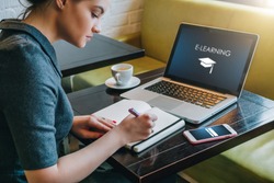 Young businesswoman sitting at table in cafe in front of laptop with inscription on screen e-learning and image of square academic cap and making notes in notebook,diary. Online education,e-learning.