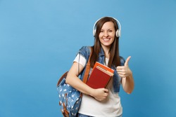 Young laughing woman student in denim clothes with backpack, headphones listening music, holding school books showing thumb up isolated on blue background. Education in high school university college