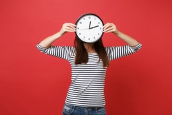 Portrait of young woman in striped clothes hiding, covering face with round clock isolated on red wall background. Time is running out. People sincere emotions, lifestyle concept. Mock up copy space