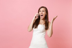 Angry Irritated bride woman in white wedding dress spreading hands, talking on mobile phone, screaming and swearing isolated on pastel pink background. Wedding to do list. Organization of celebration