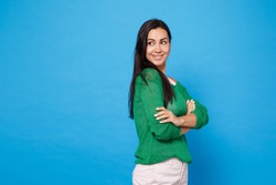 Side view of attractive smiling young woman in green casual clothes looking aside holding hands crossed isolated on bright blue wall background in studio. People lifestyle concept. Mock up copy space