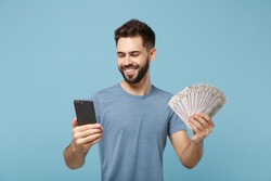 Young joyful man in casual clothes posing isolated on blue wall background, studio portrait. People lifestyle concept. Mock up copy space. Holding mobile phone, fan of cash money in dollar banknotes