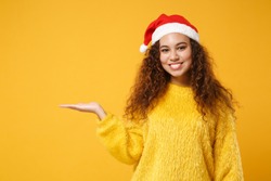 Smiling young african american Santa girl in fur sweater, Christmas hat isolated on yellow wall background. Happy New Year 2020 celebration holiday concept. Mock up copy space. Pointing hand aside