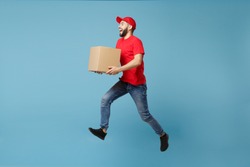 Delivery man in red uniform isolated on blue background, studio portrait. Male employee in cap t-shirt print working as courier dealer hold empty cardboard box. Service concept. Mock up copy space