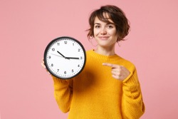 Smiling young brunette woman girl in yellow sweater posing isolated on pastel pink background in studio. People sincere emotions lifestyle concept. Mock up copy space. Pointing index finger on clock