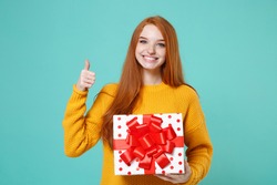 Smiling redhead girl in yellow sweater posing isolated on blue turquoise background. Valentine's Day Women's Day, birthday concept. Hold white red present box with gift ribbon bow, showing thumb up