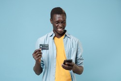 Smiling young african american man guy in casual shirt, yellow t-shirt posing isolated on blue wall background. People lifestyle concept. Mock up copy space. Using mobile phone, hold credit bank card