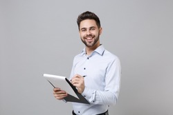 Cheerful young unshaven business man in light shirt posing isolated on grey background. Achievement career wealth business concept. Mock up copy space. Hold clipboard with papers document write notes