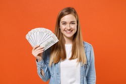 Smiling young woman girl in casual denim clothes posing isolated on orange wall background studio portrait. People lifestyle concept. Mock up copy space. Holding fan of cash money in dollar banknotes