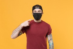 Young tattooed man guy in casual t-shirt cap posing isolated on yellow wall background studio portrait. People emotions lifestyle concept. Mock up copy space. Pointing index finger on black face mask