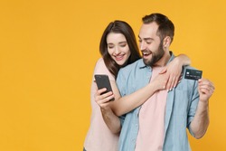 Funny young couple two friends guy girl in pastel blue casual clothes isolated on yellow background. People lifestyle concept. Mock up copy space. Hugging, using mobile phone, hold credit bank card