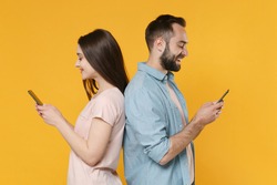 Smiling young couple friends guy girl in casual clothes isolated on yellow wall background. People lifestyle concept. Mock up copy space. Standing back to back using mobile phone typing sms message