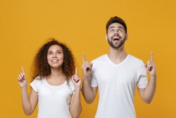 Excited young couple two friends european guy african american girl in white t-shirts isolated on yellow background in studio. People lifestyle concept. Mock up copy space. Pointing index fingers up