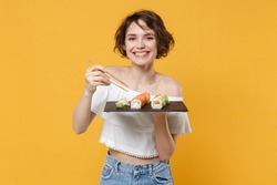 Young brunette woman girl in casual clothes hold in hand makizushi sushi roll served on black plate traditional japanese food isolated on yellow background studio portrait. People lifestyle concept