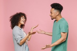 Side view of cheerful excited young african american couple two friends man woman in gray green casual clothes spreading hands speaking talking isolated on pastel pink color background studio portrait