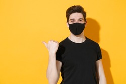 Smiling young man in black t-shirt sterile face mask to safe from coronavirus virus covid-19 during pandemic quarantine pointing finger aside workspace isolated on yellow background studio portrait.