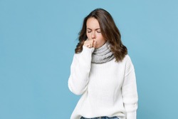 Sick young woman in white sweater gray scarf coughing covering mouth with hand keeping eyes closed isolated on blue background studio. Healthy lifestyle ill sick disease treatment cold season concept