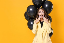 Amazed young woman in suit jacket celebrating birthday holiday party with bunch of air ballons keeping mouth open put hands on cheeks isolated on yellow background studio portrait. Black friday sale