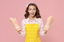 Surprised excited cheerful young brunette woman housewife 20s wearing yellow apron gloves potholders doing housework isolated on pastel pink colour background studio portrait. Housekeeping concept