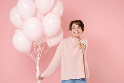 Smiling happy young brunette woman 20s in casual sweater holding bunch of air balloons, celebrating birthday holiday party, pointing index finger on camera isolated on pastel pink background studio 