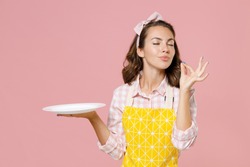 Cute young woman housewife in yellow apron hold empty plate making okay taste delight sign keeping eyes closed doing housework isolated on pastel pink background studio portrait. Housekeeping concept