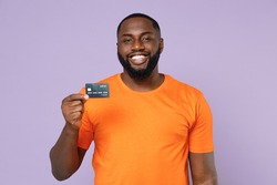Smiling funny young african american man 20s wearing basic casual orange blank empty t-shirt standing hold credit bank card looking camera isolated on pastel violet colour background studio portrait