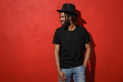 Smiling handsome attractive young african american man guy with dreadlocks 20s wearing black casual t-shirt hat posing looking aside isolated on bright red color wall background studio portrait