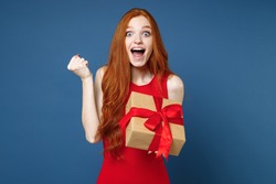 Amazed young redhead girl in red elegant evening dress hold present box with gift ribbon bow doing winner gesture isolated on blue background. St. Valentine's Day Women's Day birthday holiday concept