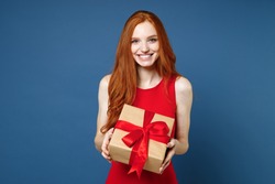Smiling young redhead woman 20s in red elegant evening dress hold present box with gift ribbon bow isolated on blue background. St. Valentine's Day International Women's Day birthday, holiday concept