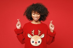 Little african american curly kid girl 13 years old in knitted cozy deer Christmas sweater keep fingers crossed isolated on bright red background children studio portrait. Childhood lifestyle concept