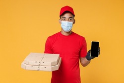 Delivery employee african man in red cap blank print t-shirt face mask gloves work courier service on quarantine coronavirus covid19 virus hold pizza flatbox mobile phone isolated on yellow background