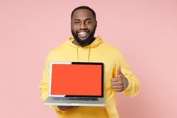 Smiling young african american man 20s wearing casual yellow streetwear hoodie hold laptop pc computer with blank empty screen showing thumb up isolated on pastel pink wall background studio portrait