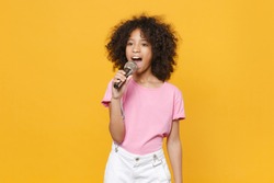 Charming little african american kid girl 12-13 years old in pink t-shirt isolated on yellow wall background studio portrait. Childhood lifestyle concept. Mock up copy space. Sing song in microphone