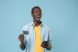Excited young african american man 20s in casual shirt, yellow t-shirt isolated on blue background. People lifestyle concept. Mock up copy space. Using mobile phone hold credit bank card looking up