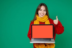 Funny young brunette woman 20s in basic knitted red sweater yellow scarf hold laptop pc computer with blank empty screen showing thumb up isolated on bright green color background studio portrait