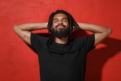 Relaxed calm smiling young african american man with dreadlocks 20s wearing black basic casual t-shirt posing sleep with hands behind head isolated on bright red color wall background studio portrait