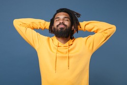 Relaxed young african american man with dreadlocks in yellow streetwear hoodie posing isolated on blue wall background studio portrait. People emotions lifestyle concept. Sleep with hands behind head