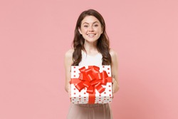 Smiling young brunette woman 20s in light casual clothes posing isolated on pastel pink wall background. Birthday holiday concept. Mock up copy space. Hold white red present box with gift ribbon bow