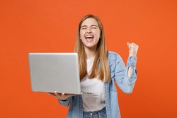 Happy young woman in casual denim clothes posing isolated on orange background studio portrait. People lifestyle concept. Mock up copy space. Hold laptop pc computer, doing winner gesture celebrating