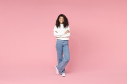 Full length young smiling african american woman 20s curly hair wear white casual knitted sweater jeans looking camera hold hands crossed folded isolated on pastel pink background studio portrait