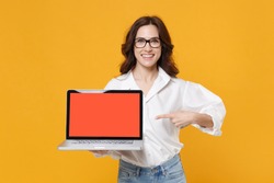Smiling young business woman in white shirt glasses isolated on yellow background studio. Achievement career wealth business concept. Point index finger on laptop pc computer with blank empty screen