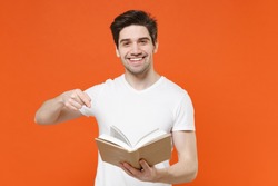 Smiling funny young man 20s wearing basic casual white blank empty t-shirt standing reading pointing index finger on book looking camera isolated on bright orange colour background, studio portrait