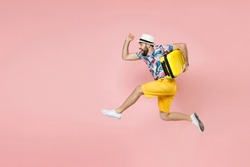 Full length side view of cheerful traveler tourist man in summer clothes hat jumping running hold suitcase isolated on pink background. Passenger traveling on weekend. Air flight journey concept