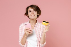 Pensive pretty young brunette woman 20s wearing casual checkered shirt standing using mobile cell phone hold credit bank card looking up isolated on pastel pink colour background, studio portrait