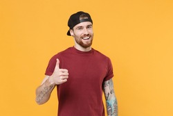 Smiling happy young bearded tattooed man 20s in casual t-shirt, black cap posing showing thumb up isolated on yellow wall background studio portrait. Mock up copy space. Tattoo translate life is fight